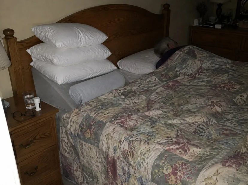 A bed at night. One side has a foam wedge, three pillows and a rolled towel. On the other side a man is asleep on one pillow.